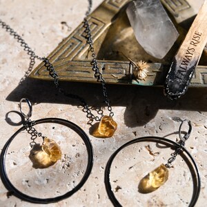 May include: A set of gold gemstone earrings with a black chain and hoop design. The earrings are on a light brown surface with a brass tray and a wooden stick with the text "Always Rise" on it.