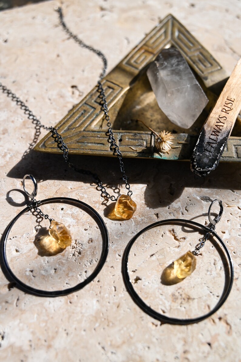 May include: A set of gold gemstone earrings with a black chain and hoop design. The earrings are on a light brown surface with a brass tray and a wooden stick with the text "Always Rise" on it.