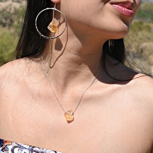 May include: A silver necklace with a raw, light orange gemstone pendant. The necklace is worn by a person with long dark hair and a silver hoop earring with a similar gemstone dangling from it.