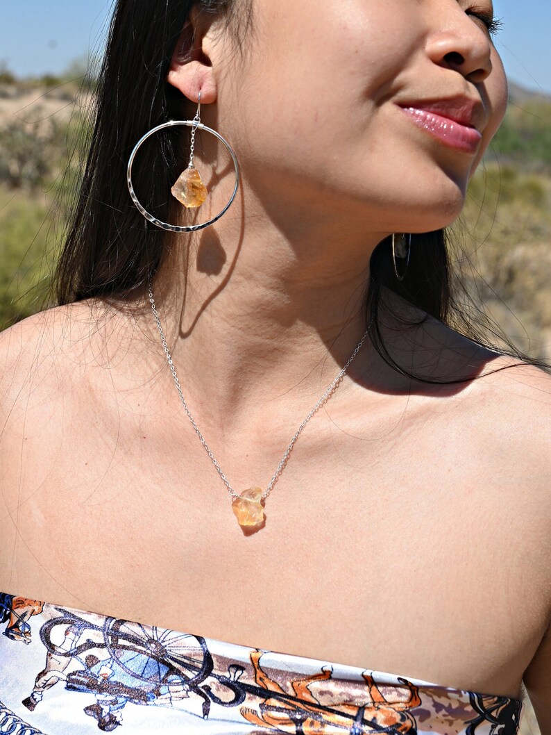 May include: A silver necklace with a raw, light orange gemstone pendant. The necklace is worn by a person with long dark hair and a silver hoop earring with a similar gemstone dangling from it.