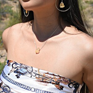 May include: A silver necklace with a yellow citrine pendant. The necklace is worn by a person with long black hair and a white and brown patterned top.