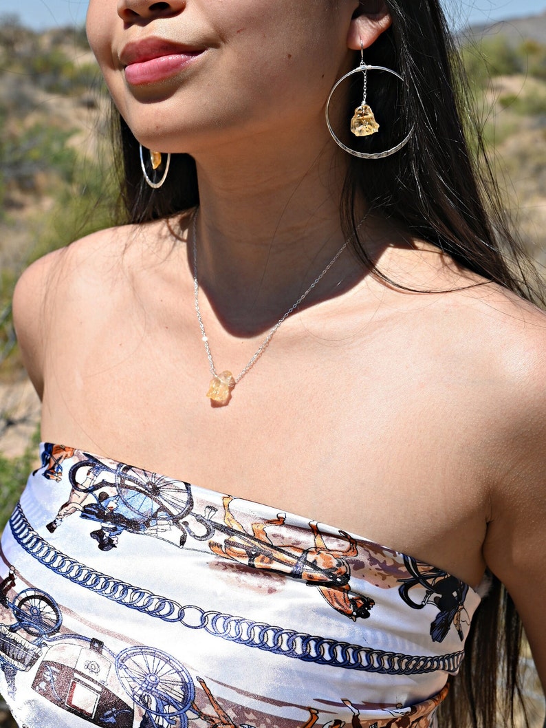 May include: A silver necklace with a yellow citrine pendant. The necklace is worn by a person with long black hair and a white and brown patterned top.