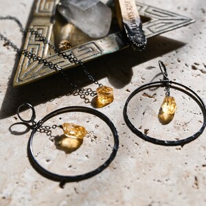 May include: A set of three jewelry pieces featuring a raw citrine stone. The set includes a necklace, a pair of earrings, and a small pendant. The jewelry is made with black metal and has a minimalist design.