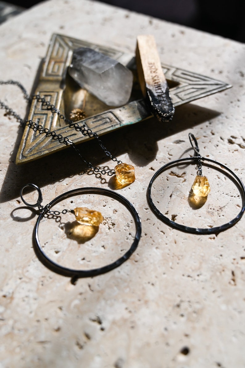 May include: A set of three jewelry pieces featuring a raw citrine stone. The set includes a necklace, a pair of earrings, and a small pendant. The jewelry is made with black metal and has a minimalist design.