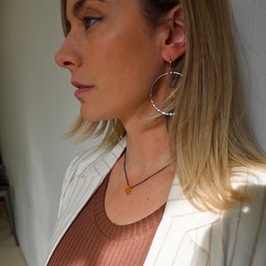 May include: A woman wearing a white blazer with gold stripes over a brown ribbed tank top. She is wearing a silver necklace with a small amber pendant and silver hoop earrings with a small amber pendant.