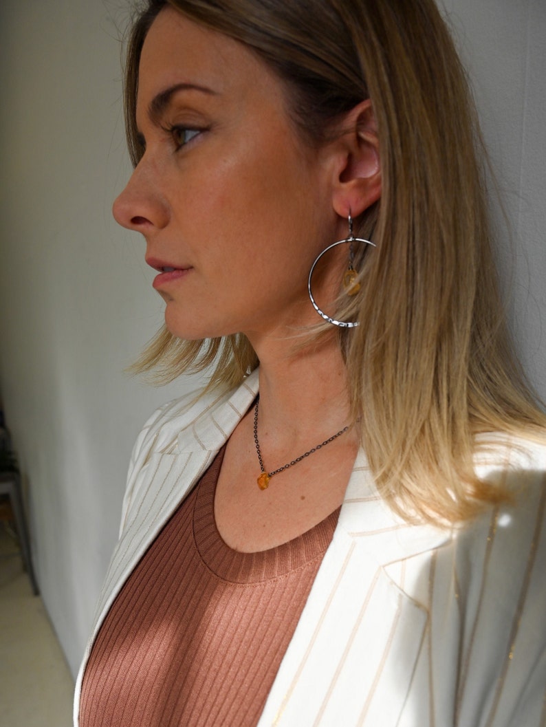 May include: A woman wearing a white blazer with gold stripes over a brown ribbed tank top. She is wearing a silver necklace with a small amber pendant and silver hoop earrings with a small amber pendant.
