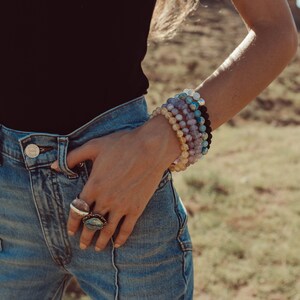 May include: A woman wearing a black tank top and blue jeans with multiple gemstone bracelets on her wrist and two rings on her fingers.