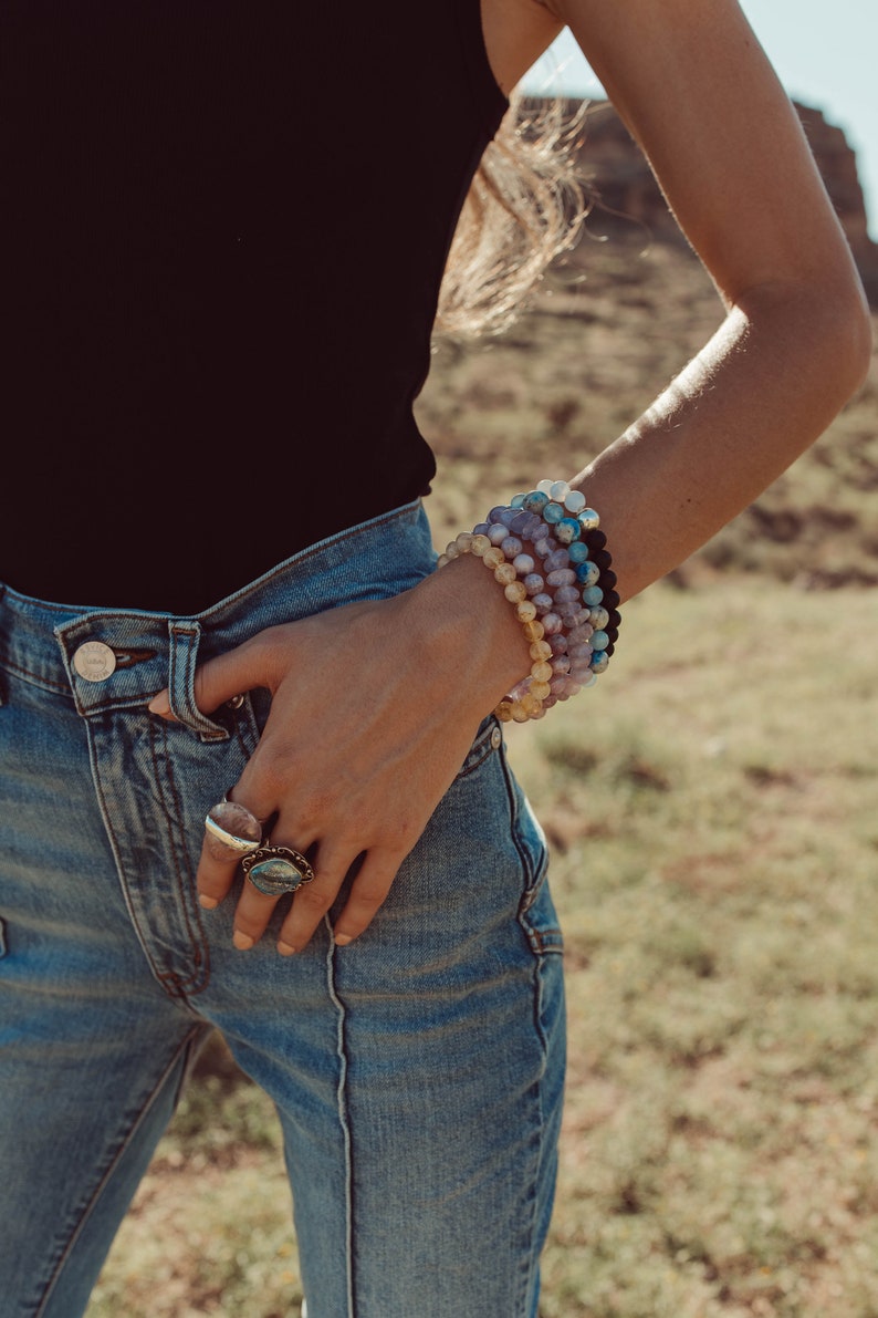 May include: A woman wearing a black tank top and blue jeans with multiple gemstone bracelets on her wrist and two rings on her fingers.