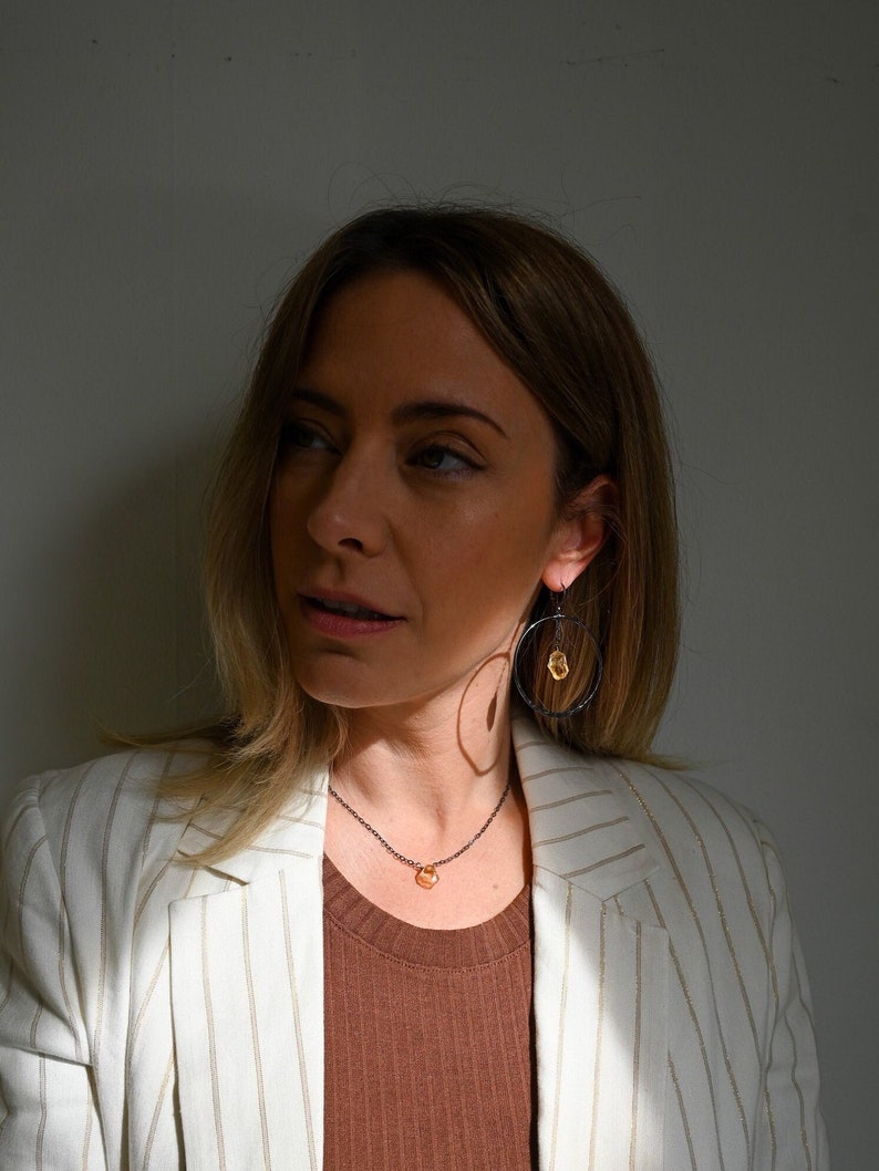 May include: A woman wearing a white striped blazer with a brown top and a delicate gold necklace with a small orange stone pendant. She is also wearing black hoop earrings with a small orange stone accent.