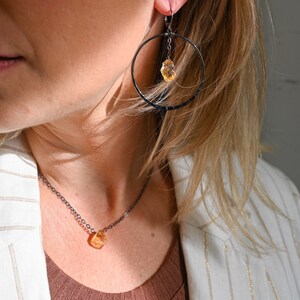 May include: A woman wearing a white blazer with gold stripes and a brown top, with a black chain necklace and matching earrings featuring a dangling orange gemstone.