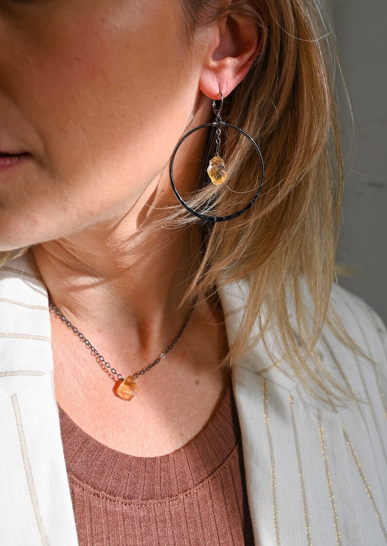 May include: A woman wearing a white blazer with gold stripes and a brown top, with a black chain necklace and matching earrings featuring a dangling orange gemstone.