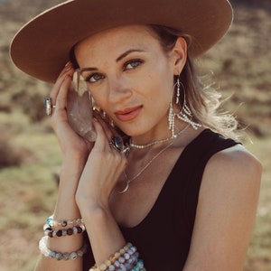 May include: A woman wearing a brown hat, a black tank top, and a silver necklace with a crescent moon pendant. She is holding a clear crystal in her hand and wearing multiple bracelets on her wrist.