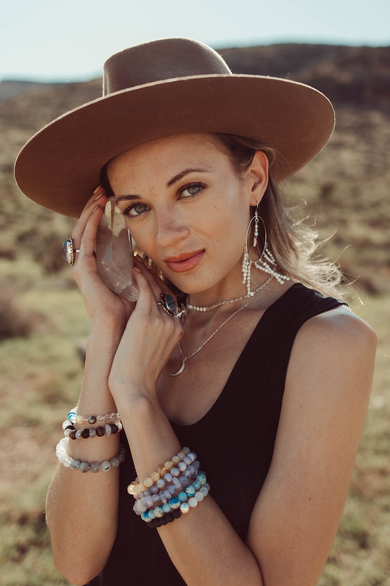 May include: A woman wearing a brown hat, a black tank top, and a silver necklace with a crescent moon pendant. She is holding a clear crystal in her hand and wearing multiple bracelets on her wrist.
