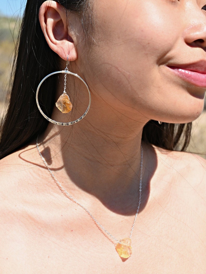 May include: A silver necklace and earrings with a raw, yellow-orange gemstone pendant and drop. The necklace is a delicate chain with a small, raw gemstone pendant. The earrings are hoop earrings with a raw gemstone dangling from the hoop.