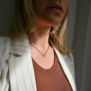 May include: A woman wearing a white blazer with gold stripes over a brown ribbed tank top. She is wearing a delicate gold necklace with a small, faceted, amber-colored stone pendant.
