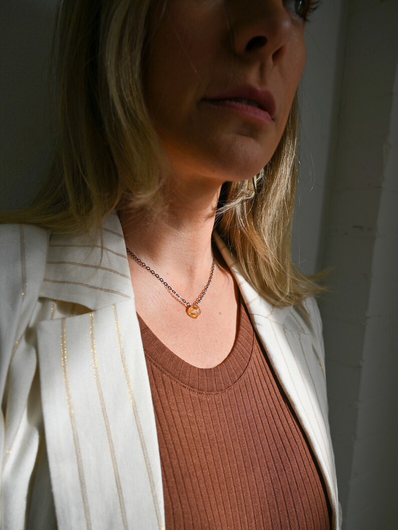 May include: A woman wearing a white blazer with gold stripes over a brown ribbed tank top. She is wearing a delicate gold necklace with a small, faceted, amber-colored stone pendant.