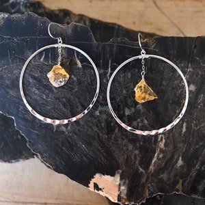 May include: A pair of silver hoop earrings with a hammered texture. Each earring features a dangling, irregular-shaped, yellow gemstone.