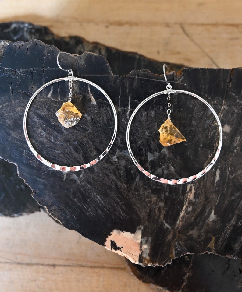 May include: A pair of silver hoop earrings with a hammered texture. Each earring features a dangling, irregular-shaped, yellow gemstone.