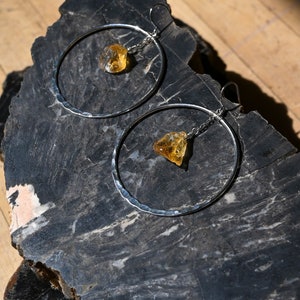 May include: A pair of silver hoop earrings with a hammered texture. Each earring features a dangling, raw, yellow citrine gemstone.