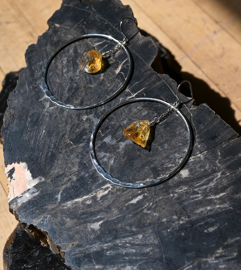 May include: A pair of silver hoop earrings with a hammered texture. Each earring features a dangling, raw, yellow citrine gemstone.