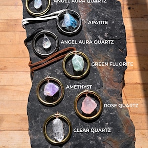 May include: Seven crystal pendants on gold and silver chains, displayed on a black slate background. The crystals are: Angel Aura Quartz, Apatite, Angel Aura Quartz, Green Fluorite, Amethyst, Rose Quartz, and Clear Quartz.