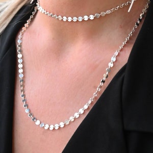 May include: Two silver necklaces with small round discs, layered on a person's neck. The necklaces are worn over a black shirt.