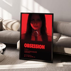 Puede incluir: Un póster de película enmarcado titulado Obsession que presenta un gran retrato en tonos rojos de una persona mirando hacia abajo a una pequeña figura en el centro. El texto incluye una definición de obsesión y la fecha, Only in theaters May 18.