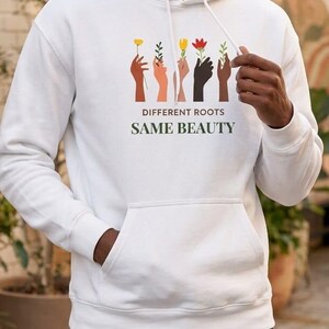 Pode incluir: Moletom branco com bolso frontal e capuz com cordão. A frente do moletom apresenta o texto "DIFFERENT ROOTS SAME BEAUTY" acima de uma ilustração de mãos segurando flores em várias cores.