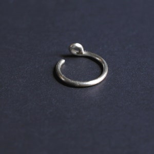 thick silver ear cuff laying on a flat black surface. One end resembles a real piercing, the other side has a curly end for comfortable wear.