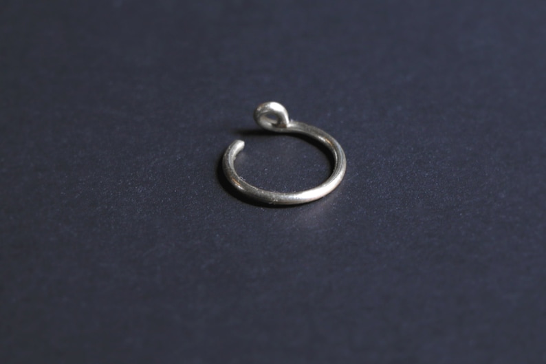 thick silver ear cuff laying on a flat black surface. One end resembles a real piercing, the other side has a curly end for comfortable wear.