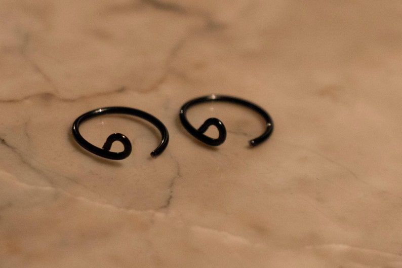 two black lip cuffs laying on a flat marble surface. One end looks like a normal piercing, the other side has a curly end for comfortable wear