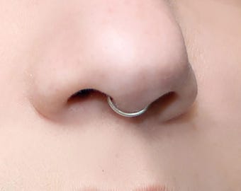 Puede incluir: Un anillo de septum circular, fino y de color plateado, colocado en la nariz. La joya es un aro de metal liso y continuo que se asienta de forma segura en el tabique nasal, con una superficie pulida, reflectante y un diseño circular.