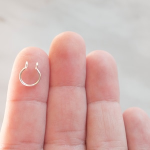 May include: A silver nose ring with a hoop design. The ring is held between two fingers.
