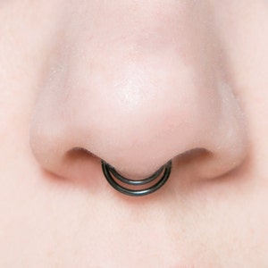 May include: Close-up of a person's nose with a black double hoop nose ring.