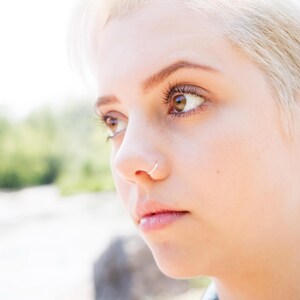 May include: Close-up portrait of a person with short, platinum blonde hair. The person has a piercing in their nose and is looking off to the side.
