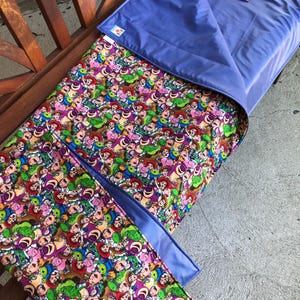May include: A rectangular nap mat featuring a colorful repeating pattern of various animated characters including cowboys, aliens, and bears. The mat has a solid periwinkle blue underside and a matching attached pillow section with a small patterned strap.