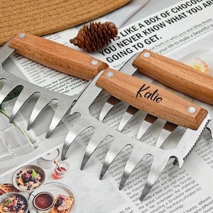 Puede incluir: Garras para carne de acero inoxidable con mangos de madera, una grabada con "Katie". Las garras están diseñadas para desmenuzar carne. La imagen también incluye una piña y un fondo de periódico.