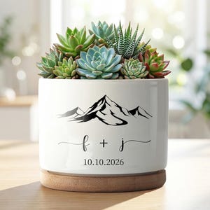 May include: A white ceramic plant pot featuring a mountain design and the initials "f + j" with the date "10.10.2026". The pot is filled with various succulents and rests on a wooden base.