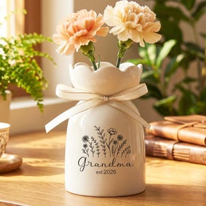 Op de afbeelding: Een witte keramische vaas met een geschulpte bovenkant, vastgebonden met een crèmekleurig lint, met twee perzikroze anjers. De vaas heeft een bloemenontwerp en het woord "Grandma" met "est. 2026" erop gedrukt.