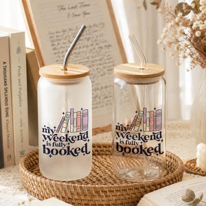May include: Two glass tumblers with bamboo lids and straws sit on a woven tray. Each tumbler has a graphic of books and the text "my weekend is fully booked." One tumbler is frosted, the other clear. Books and a handwritten note are in the background.