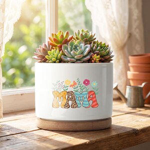 May include: A white ceramic plant pot featuring the word "MAMA" in colourful, patterned letters, with floral illustrations. The pot contains a variety of succulents. It rests on a wooden base, near a window.