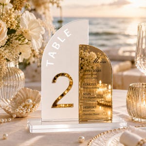 May include: A dual-panel acrylic table sign featuring a white arched section with the gold-colored number 2 and the word TABLE, alongside a gold-mirrored arched section displaying a printed menu with sections for appetizers, starters, main, and dessert.