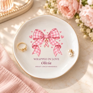 Op de afbeelding: Een ronde witte keramische schaal met een gouden rand, voorzien van een roze gingham strik, een roze bewustzijnslintje en kleine hartjes. De tekst luidt: WRAPPED IN LOVE, Olivia, BREAST CANCER AWARENESS. Een gouden ring en een parel liggen erin.