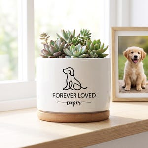 May include: A white ceramic planter with a black dog outline and the words "FOREVER LOVED cooper". The planter holds several green and purple succulents and sits on a wooden base. A framed photo of a golden retriever puppy is in the background.