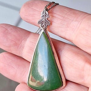 Puede incluir: Un colgante triangular con una piedra verde oscuro lisa engastada en un marco de metal plateado. El colgante está rematado con una pequeña anilla de metal plateado texturizado en forma de hoja y unido a una cadena fina de metal oscuro.