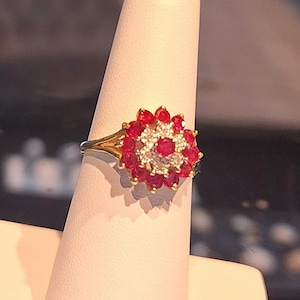 May include: A gold ring featuring a floral cluster design with a central ruby surrounded by a ring of small white stones, encircled by a larger outer layer of vibrant red rubies. The band is thin and polished, showcasing a classic prong setting.