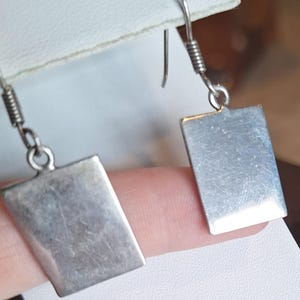 May include: A pair of silver rectangular dangle earrings. Each earring features a rectangular pendant suspended from a hook with a coiled wire detail. The earrings are shown against a neutral background.
