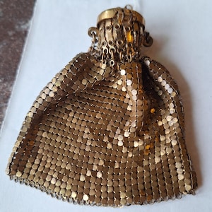 Può includere: Una pochette in maglia metallica color oro con una struttura flessibile a maglie quadrate intrecciate. La borsa presenta una cornice metallica circolare sulla parte superiore con una serie di piccoli anelli collegati lungo il bordo.