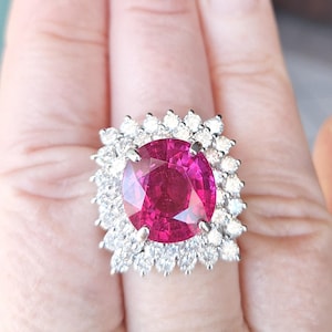 May include: A large, oval-cut, ruby-colored gemstone ring. The ruby is surrounded by a halo of smaller, clear diamonds set in a silver or white gold band. The ring is a statement piece, showcasing a vibrant, eye-catching design.