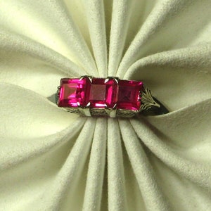 Puede incluir: Un anillo con tres piedras preciosas rosas cuadradas engastadas en una banda de plata.